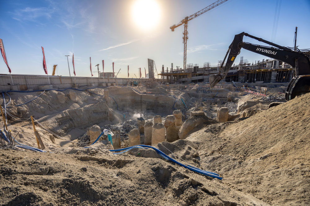Cotier House 2 By Imtiaz Construction site with a deep excavation, pillars emerging, machinery at work, under a bright sun and blue sky.